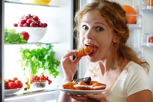 Fiatal nő a nyitott hűtő előtt állva édességet eszik